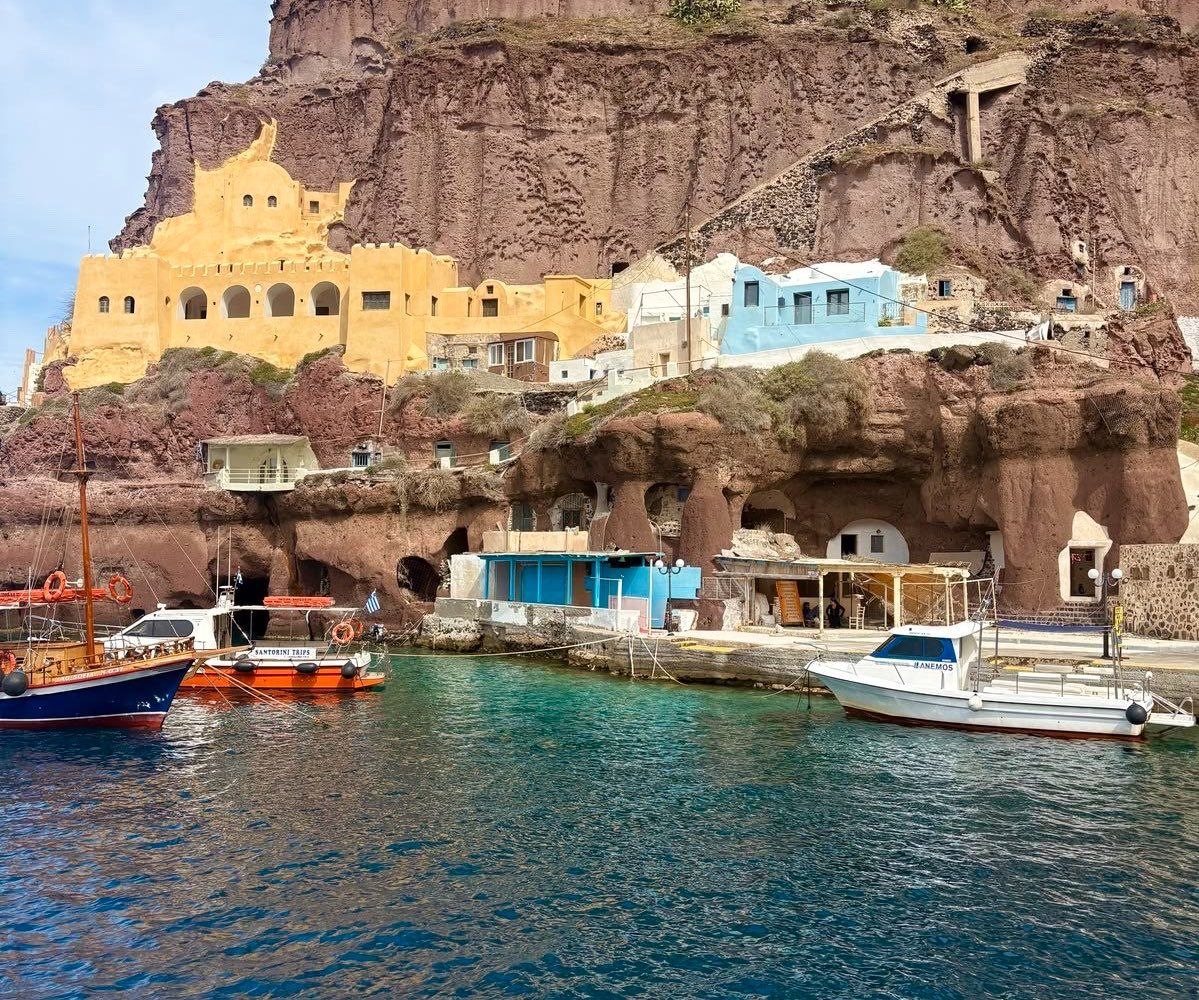 Colorful boats at Ammoudi harbor Santorini including Santorini Trips and Anemos boats with red volcanic cliffs